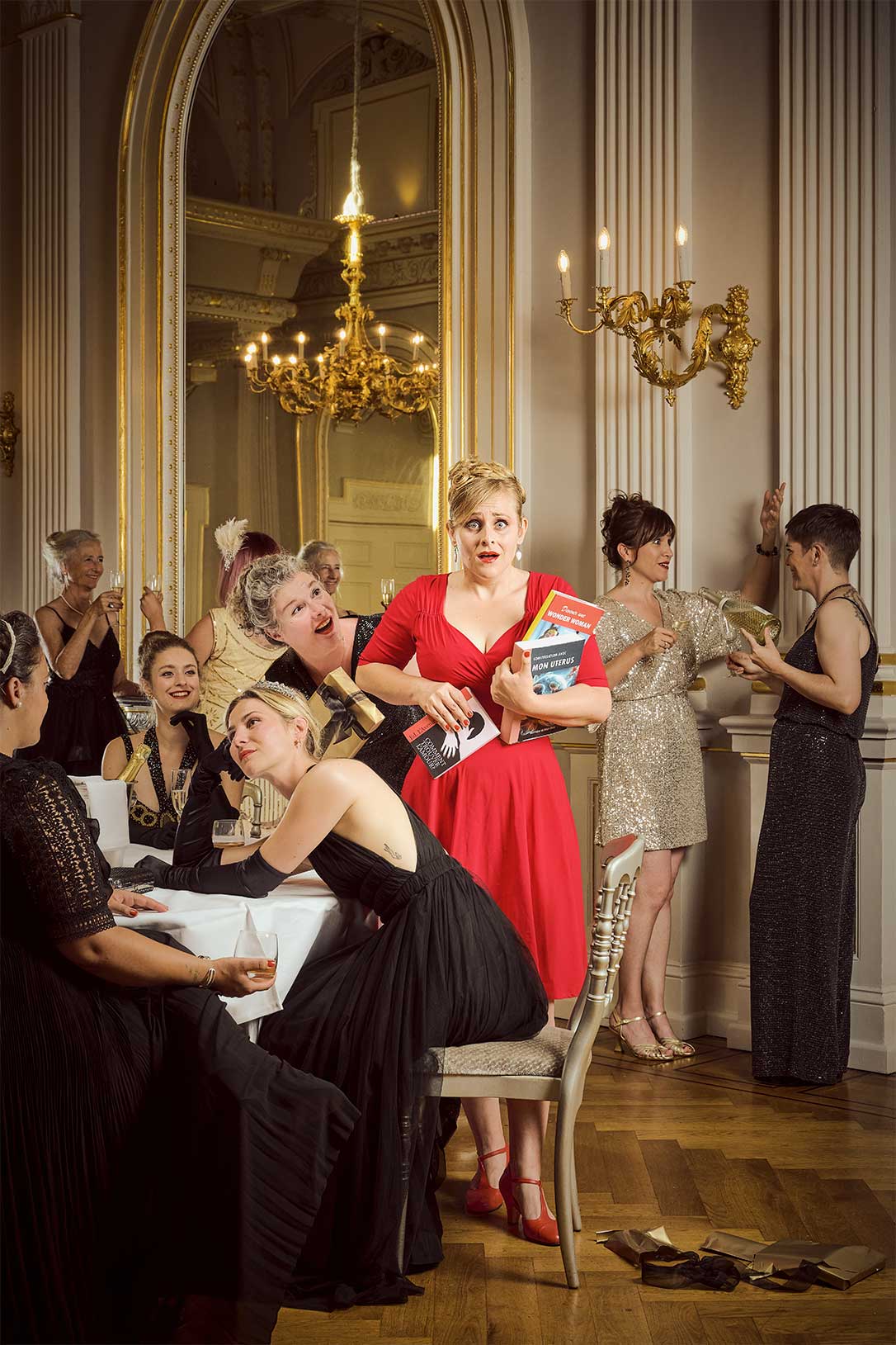 Une femme (Charlotte Thibaud-Moussouli) de 40 ans habillée en rouge tient dans ses bras des livres de développement personnel qu'elle a reçu pour son anniversaire. Son expression est celle d'une femme désemparée par la situation. Elle est entourée de femmes habillées de tenues de soirée très chics qui boivent du champagne. Elles sont dans une luxueuse salle de bal.